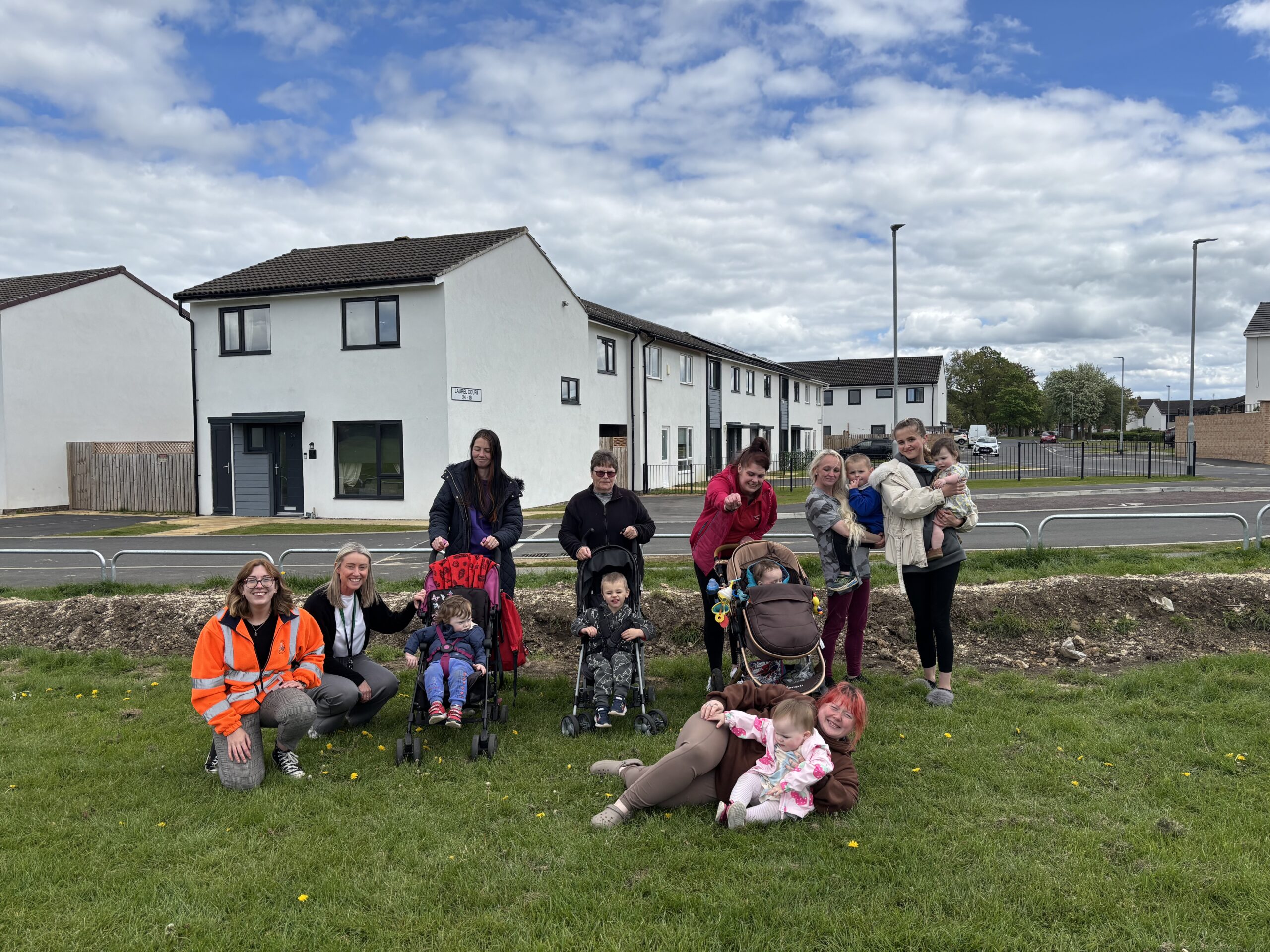 IMG_5756 A group of people, including adults and children, gather outside on a lawn in a residential area.