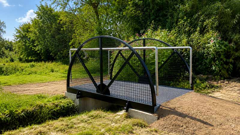 Small black metal footbridge with arched sides over a shallow ditch in a grassy area.