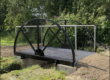 A small metal footbridge over a narrow ditch, surrounded by green grass and leafy trees.