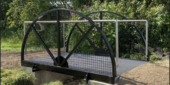 Screenshot 2026-07-02 090107 A small metal footbridge over a narrow ditch, surrounded by green grass and leafy trees.