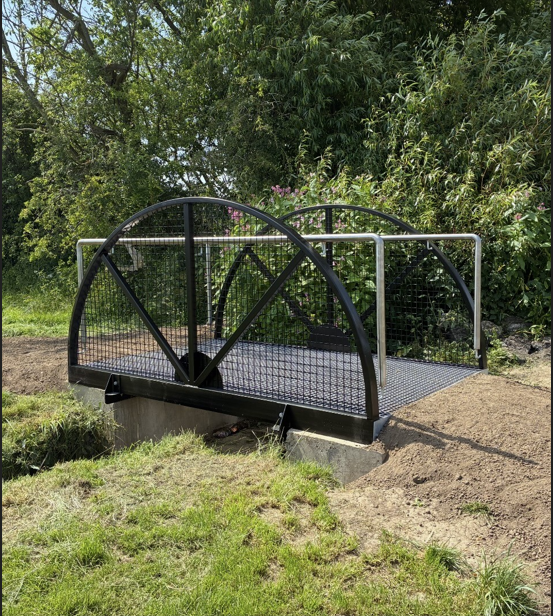 Screenshot 2026-07-02 090107 A small metal footbridge over a narrow ditch, surrounded by green grass and leafy trees.