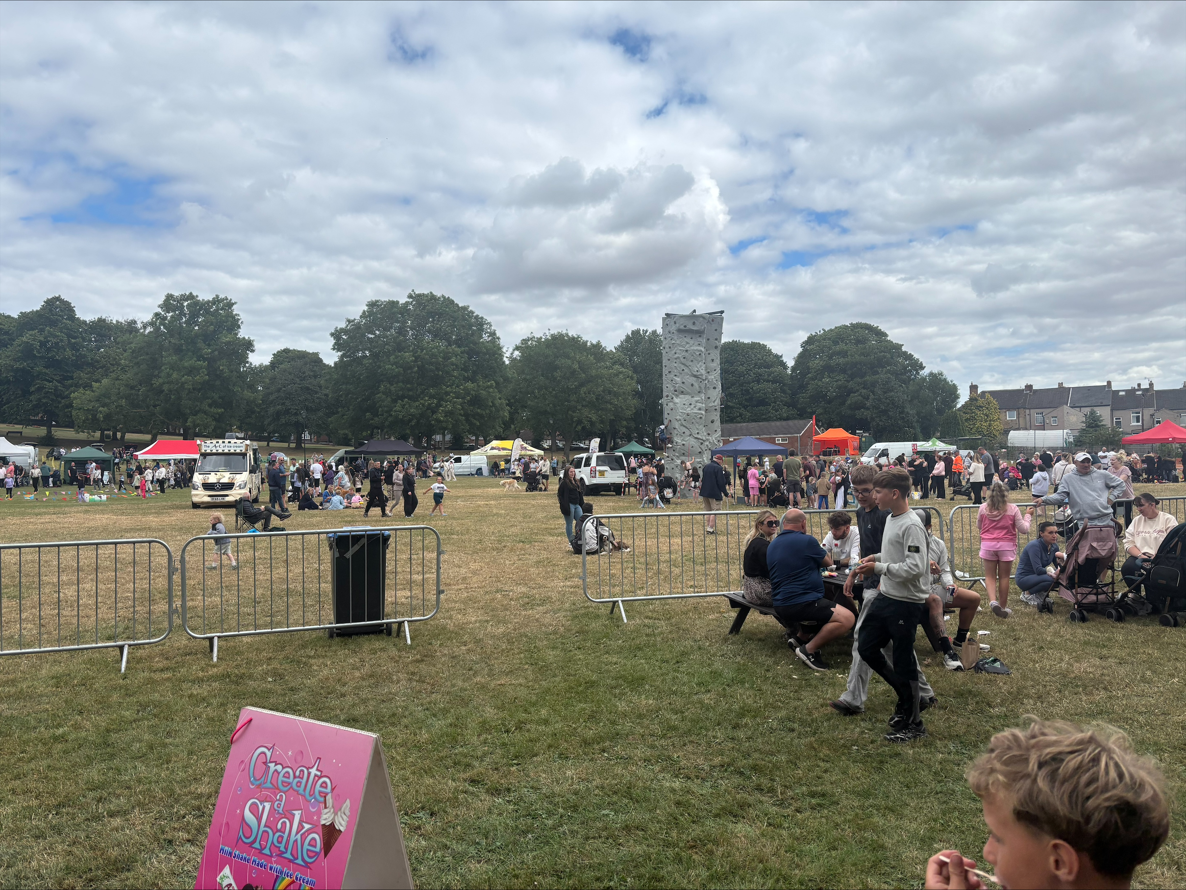 processed-A9E6D3A1-5F72-4D6E-BC40-0D2C31237C09 People at an outdoor fête with marquees, a climbing wall, and picnic tables on a grassy field.